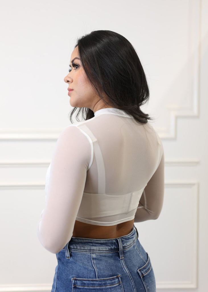 Woman in a white long-sleeve top and blue jeans standing against a white wall.