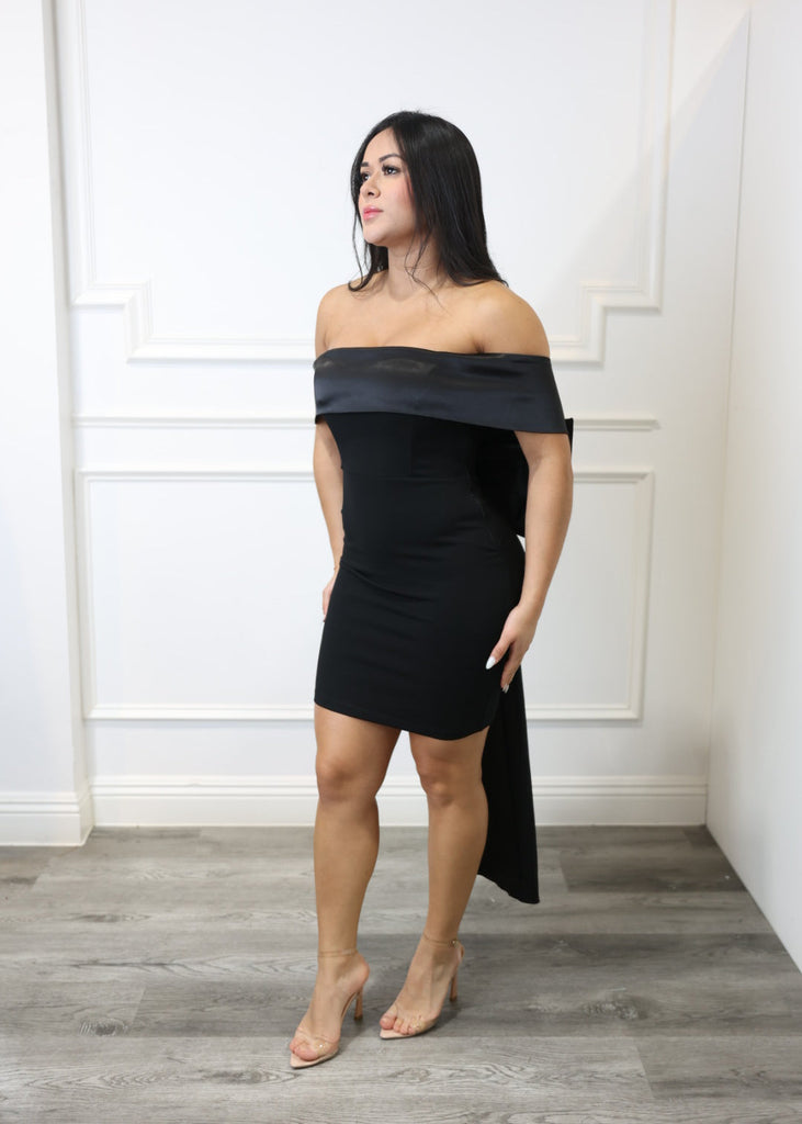 Woman in a black dress standing against a white wall with wooden floor.